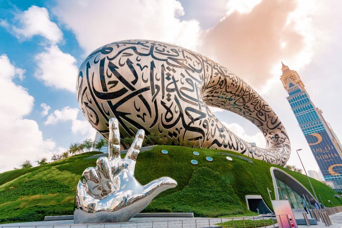 Large metallic hand sculpture with Arabic calligraphy at Museum of the Future in Dubai, with minaret tower visible in backgro
