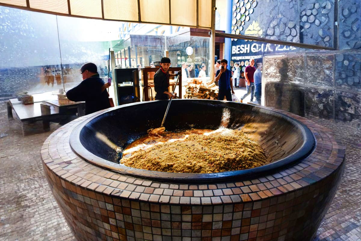 Explore Uzbekistan Large traditional Uzbek cauldron filled with plov dish at indoor market or bazaar with visitors and wooden shop stalls