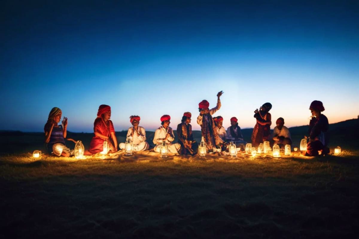Group of people sitting in circle on desert ground at dusk surrounded by glowing lanterns under blue twilight sky