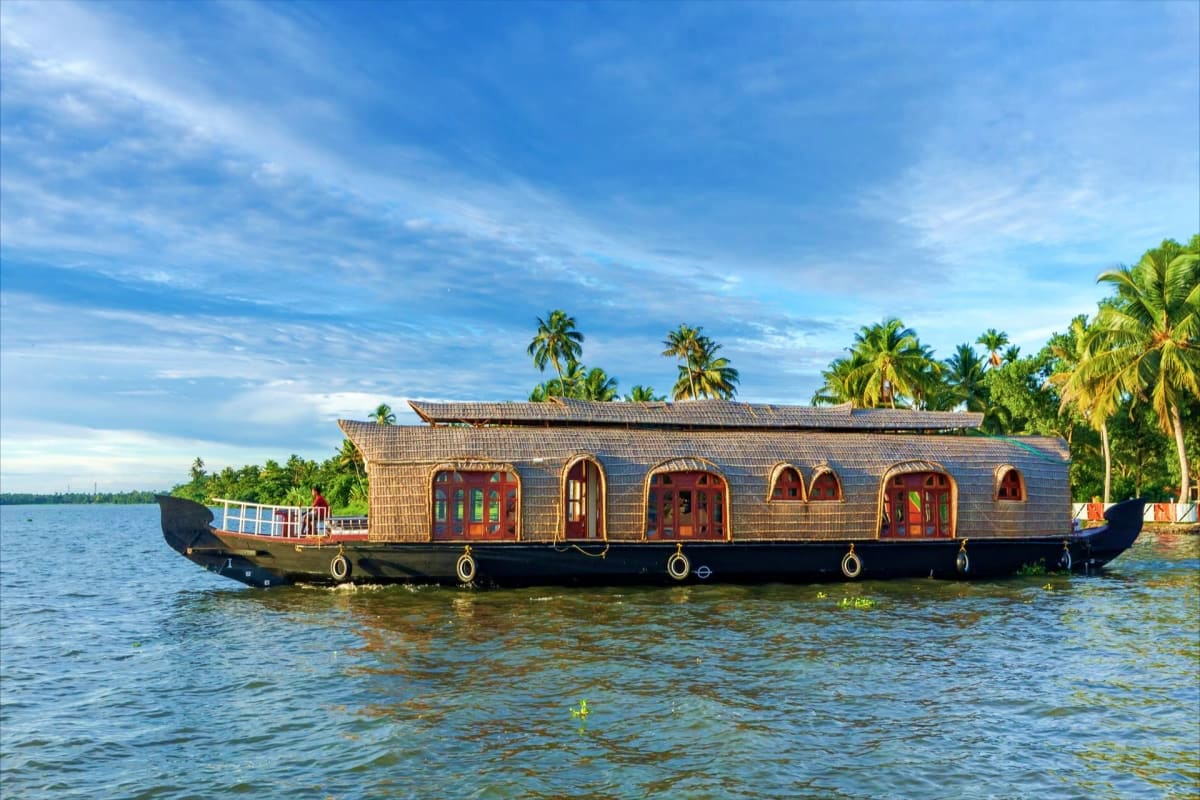 Traditional wooden houseboat with red doors floating on calm backwater waters surrounded by palm trees under blue sky