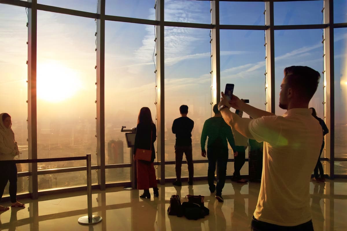 Tourists photographing sunset cityscape from high-rise observation deck with floor-to-ceiling windows overlooking Dubai skyli