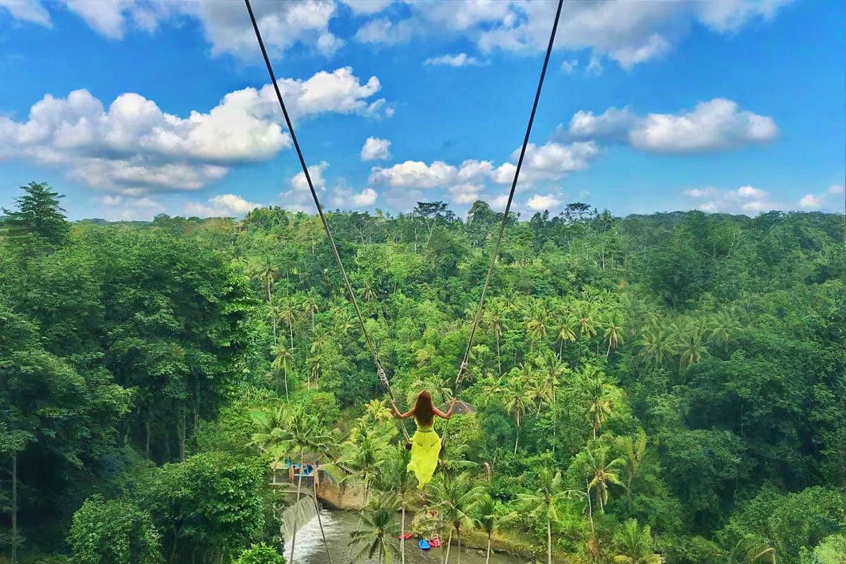 Woman on jungle swing wearing yellow dress overlooking lush rainforest canopy and river in Bali