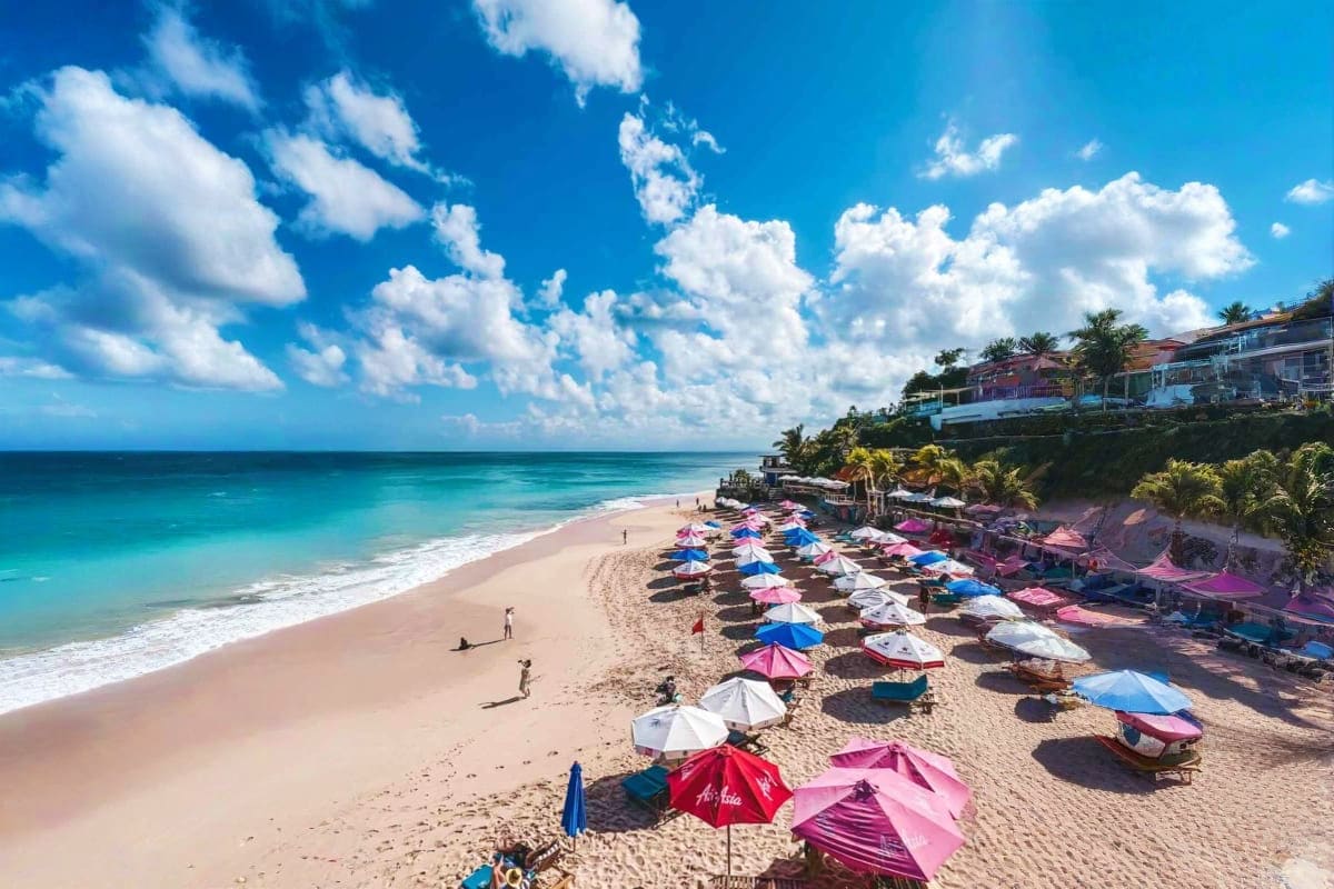 Colorful umbrellas dot a sandy beach in Bali with turquoise ocean waves, white clouds, and beachfront resort buildings on a c