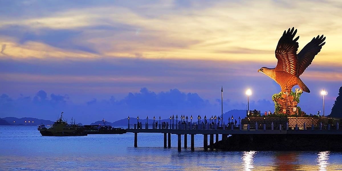 Golden eagle statue at Langkawi's waterfront pier at dusk with boats and mountains in background