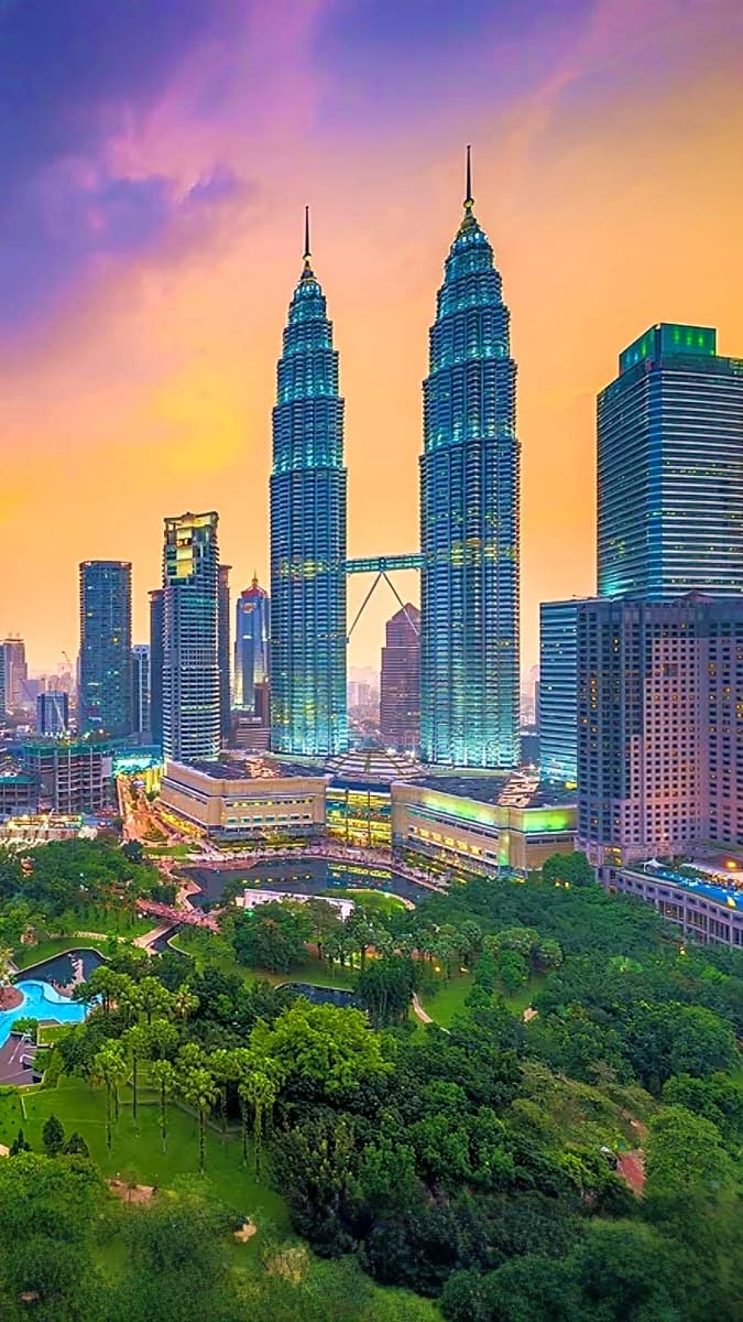 Petronas Twin Towers illuminated at sunset with Kuala Lumpur skyline and lush green park in foreground