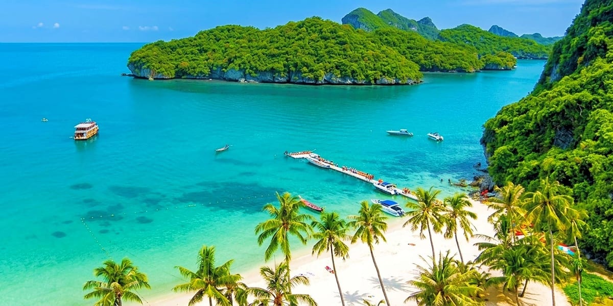 Tropical beach in Koh Samui with turquoise water, white sand, palm trees, limestone cliffs, and moored boats with forested is