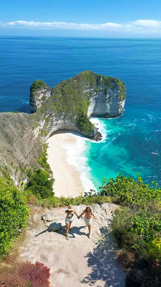 Two tourists standing on cliff overlooking Kelingking Beach with turquoise waters and limestone rock formations in Bali, Indo