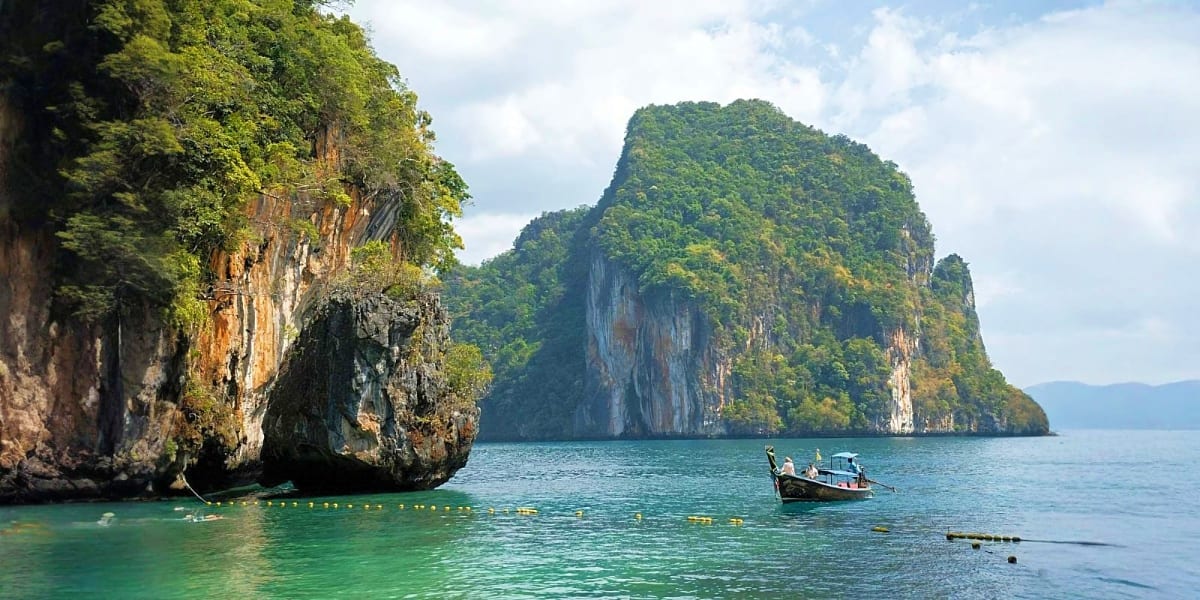 Limestone cliffs with dense vegetation and traditional longtail boat in turquoise waters of Thai islands