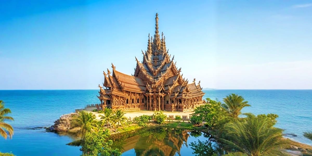 Golden Thai temple with ornate spires on rocky peninsula surrounded by turquoise ocean and palm trees under clear blue sky