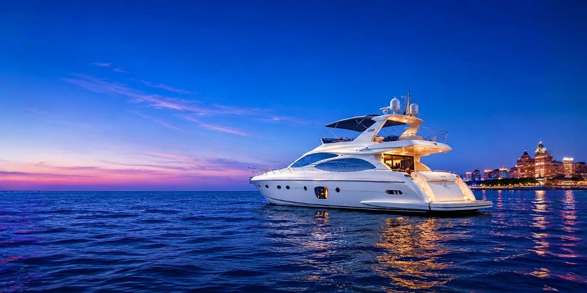 Luxury white motor yacht anchored in calm blue water at dusk with illuminated coastal city skyline in background