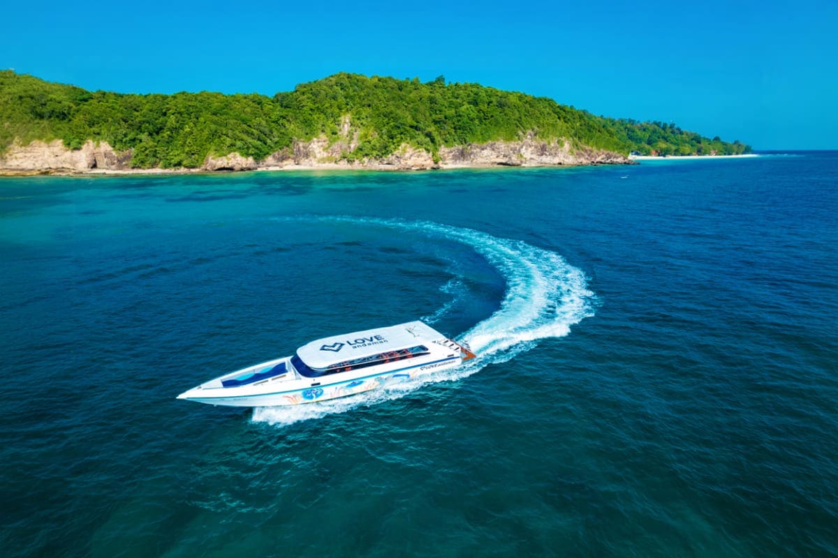 Speedboat with white wake cruising turquoise waters past forested limestone cliffs and beach during island tour