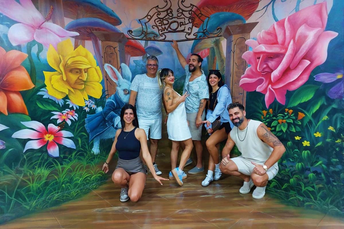 Group of six people posing together inside colorful 3D mural room with giant flowers, blue rabbits, and ornate architectural 