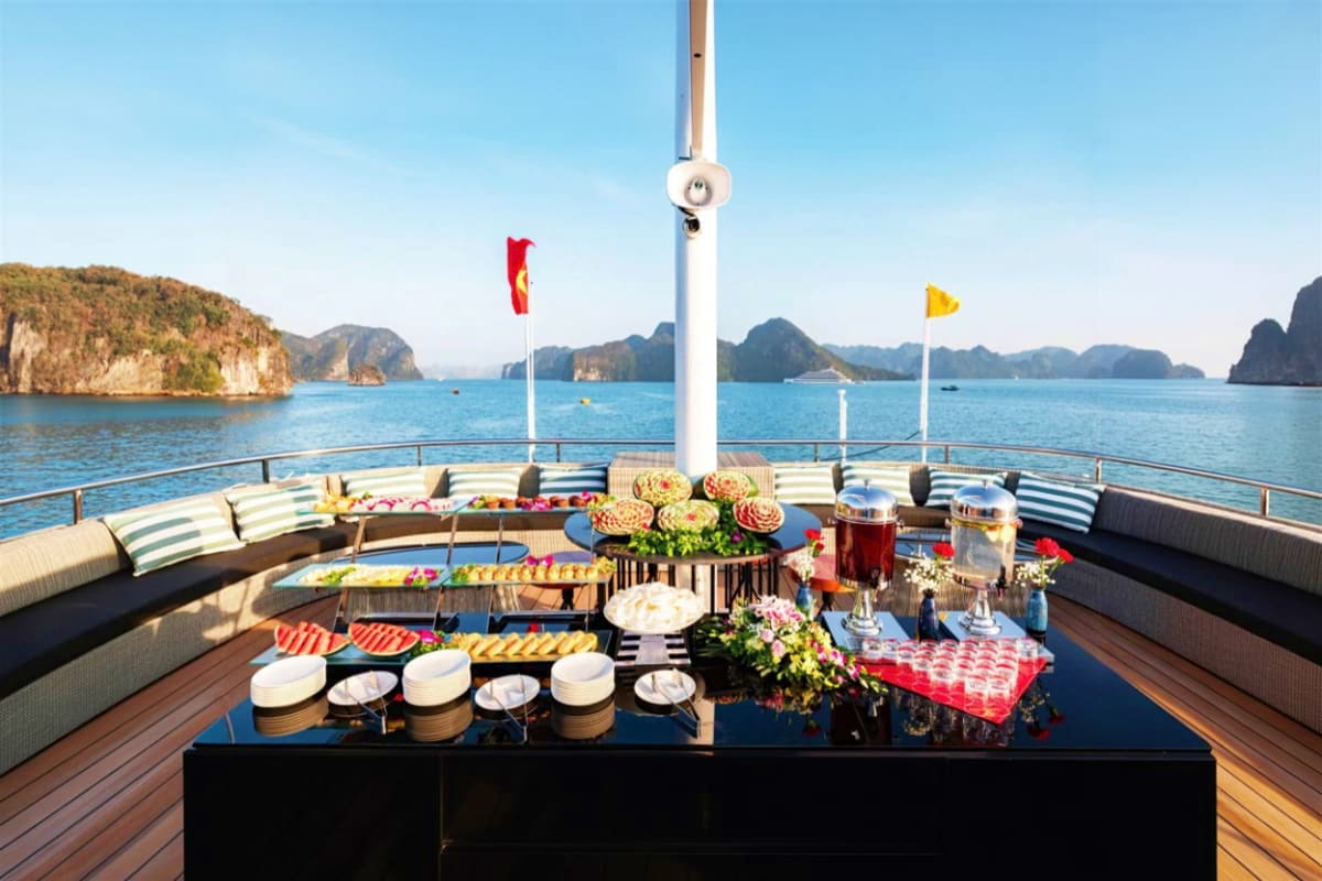 Luxury boat deck in Ha Long Bay with buffet spread, Vietnamese flags, limestone karst mountains, and turquoise waters under c