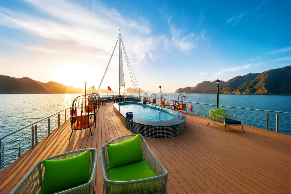 Luxury cruise ship deck with swimming pool, loungers, and sailboat on Ha Long Bay waters at sunrise with limestone mountains.