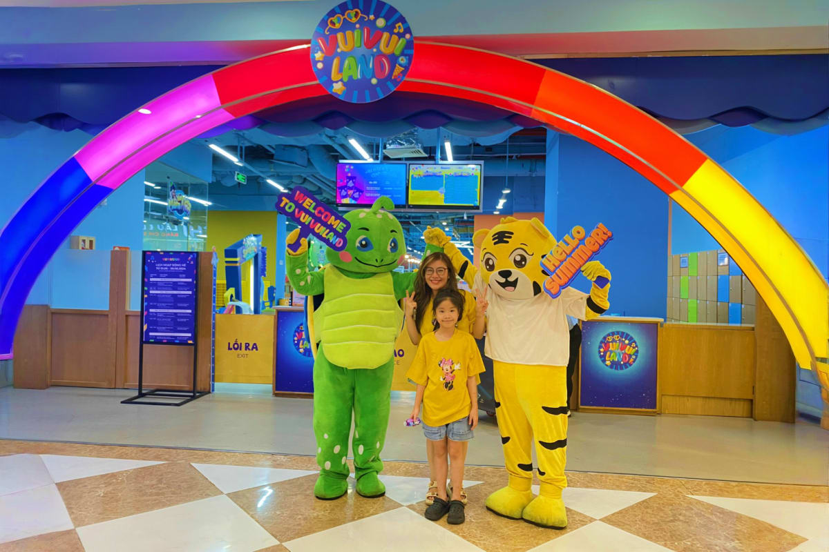 Woman and child posing with green turtle and yellow dog mascots under colorful rainbow arch at VinKE amusement park entrance