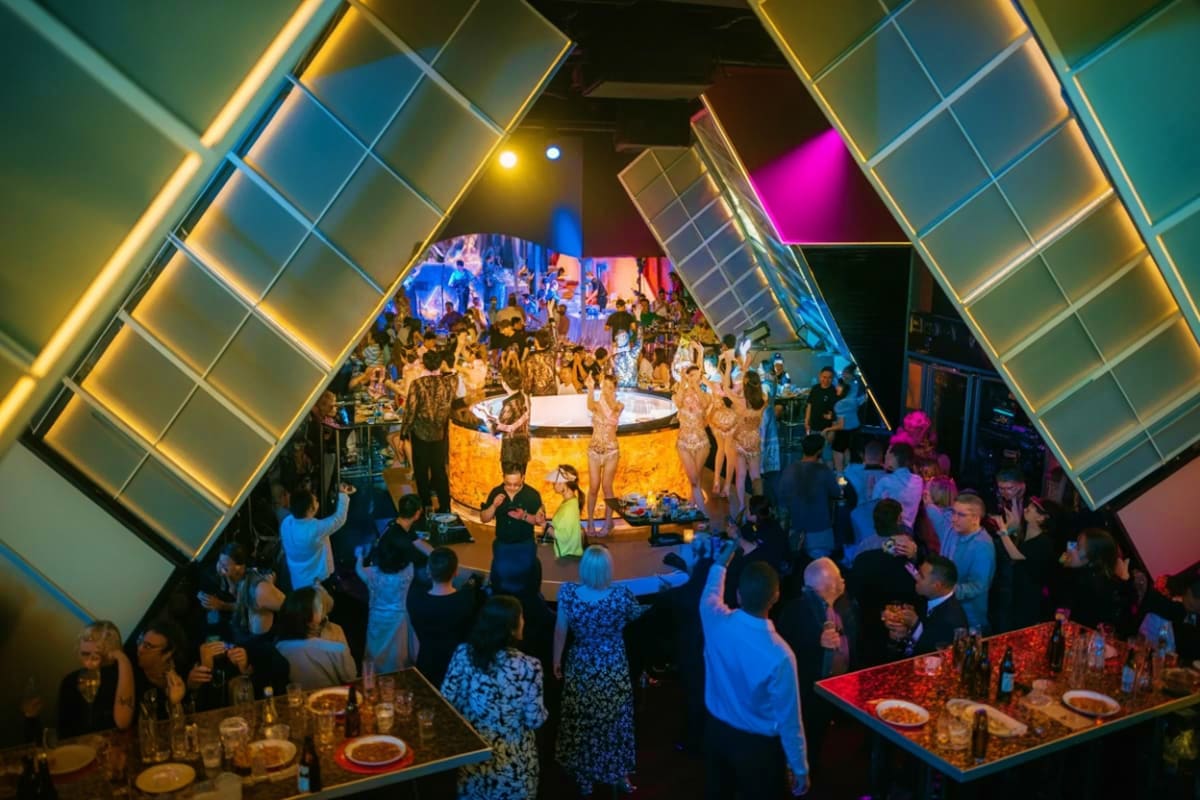 Overhead view of an upscale nightclub or dinner venue with illuminated geometric ceiling panels, dining tables, and crowds of