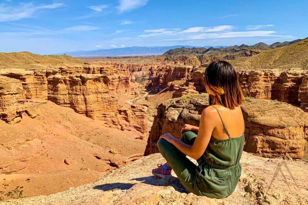 Woman sitting on rocky cliff overlooking vast canyon with layered red rock formations and mountains in distance