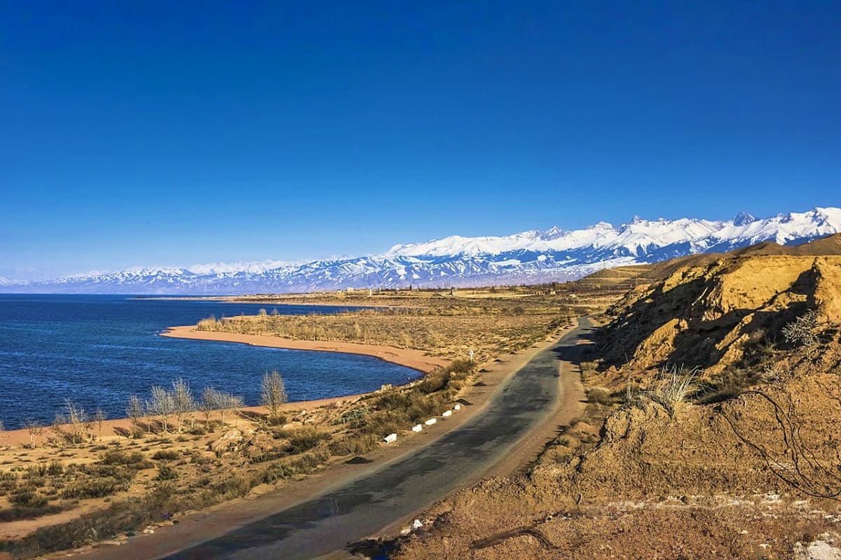 Scenic mountain lake landscape with snow-capped peaks, winding road along blue water, and desert shoreline in Kyrgyzstan