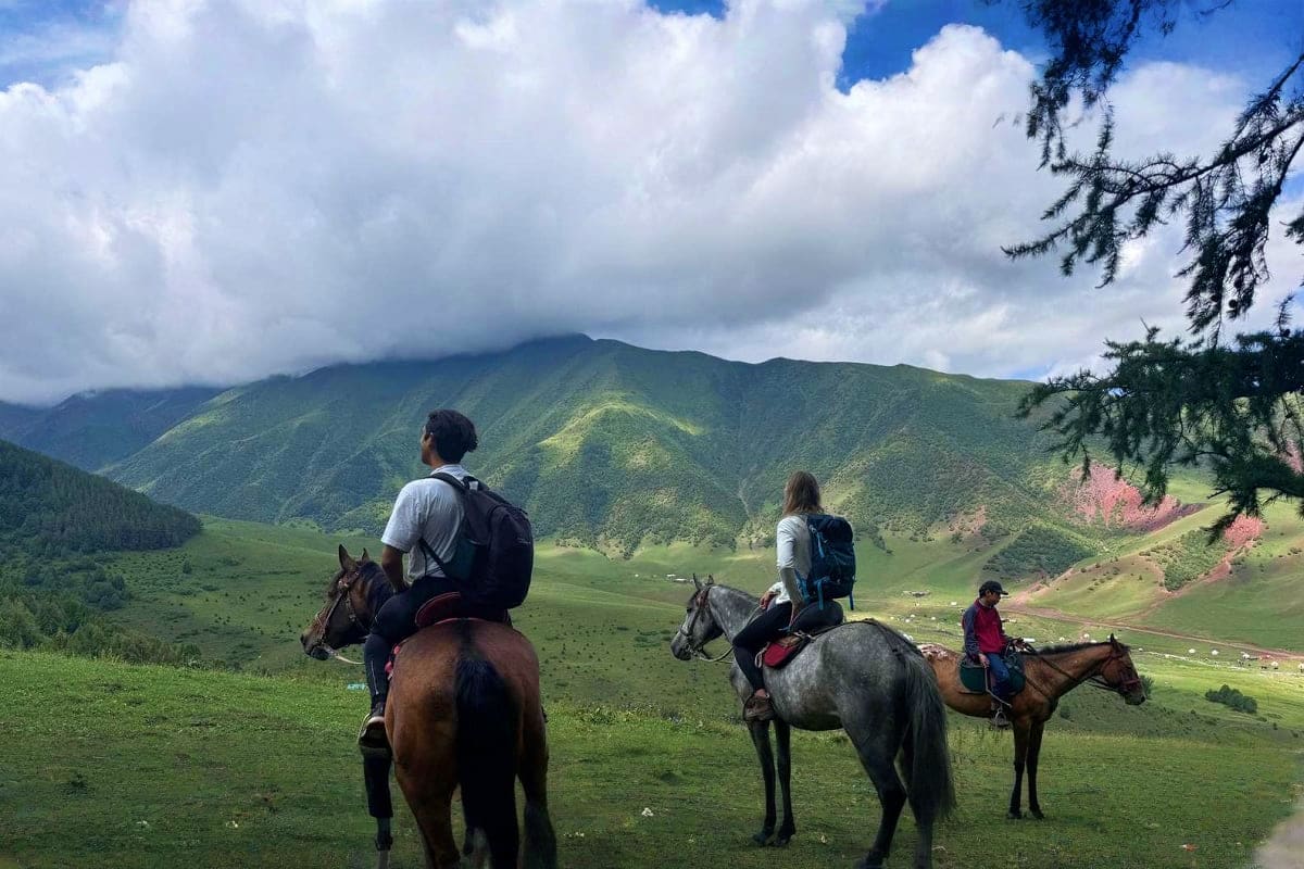 Group of tourists horseback riding in green mountain valley with forested hills and clouds in Chunkurchak