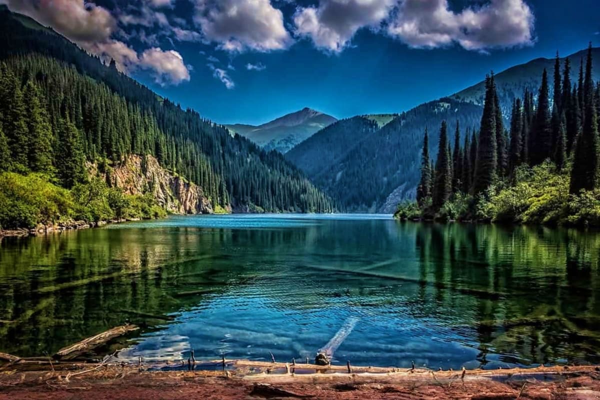 Pristine mountain lake surrounded by dense coniferous forests and towering peaks under blue sky with white clouds