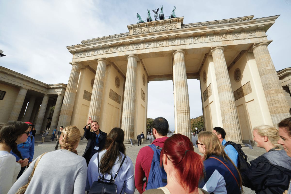 Tourist group listening to guide at Brandenburg Gate in Berlin, with massive stone columns and architectural details visible