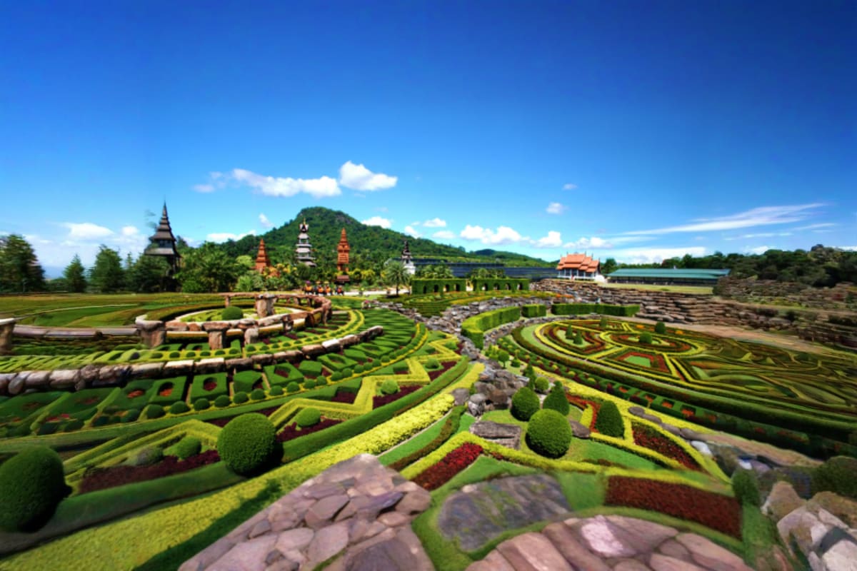 Nong Nooch Tropical Garden with ornate temples, manicured hedges, colorful flower beds, and forested hillside under blue sky