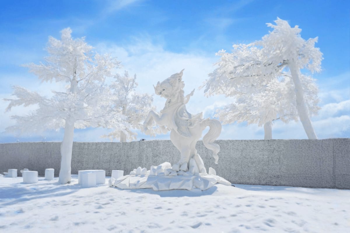 All-white winter-themed sculptural garden with frost-covered trees, white horse statue, and stone wall under blue sky at Fros