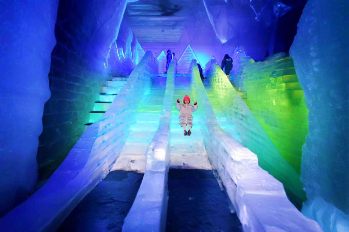 Person sledding down an icy slide in a neon-lit ice tunnel with blue and green lighting effects and other visitors in the bac