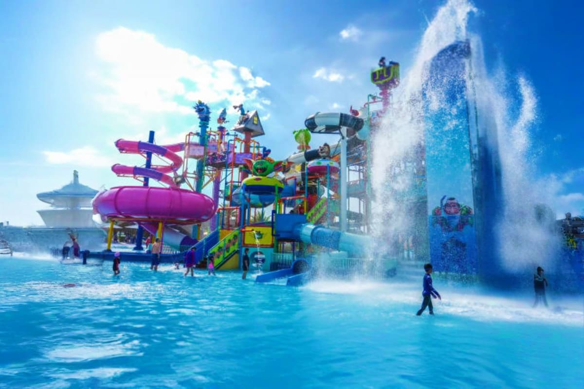 Colorful waterpark with purple slide tower, blue structure, massive water bucket dumping on guests, and children playing in b