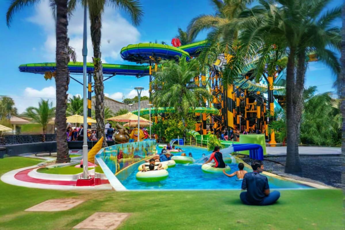 Colorful waterpark with blue lazy river, kids floating on tubes, yellow play structures, green water slides, and palm trees u