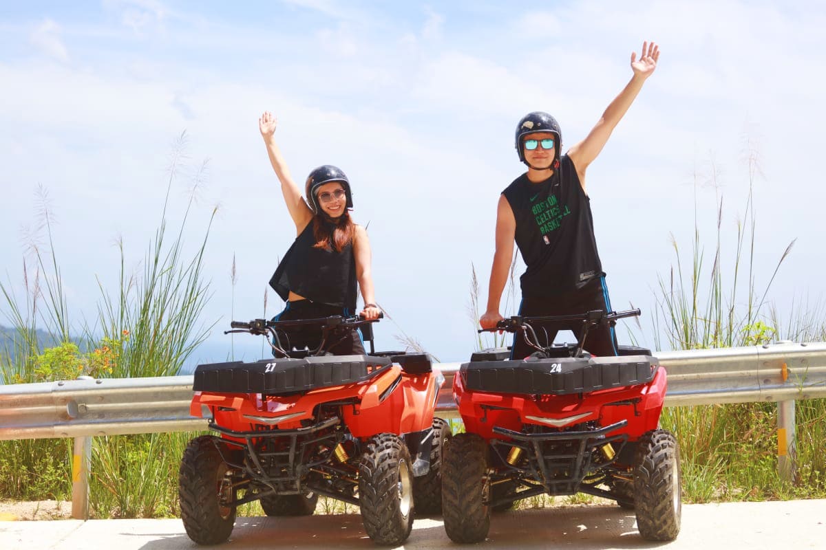 Two riders on red ATVs wearing helmets and sunglasses with arms raised, coastal landscape and dune grass in background