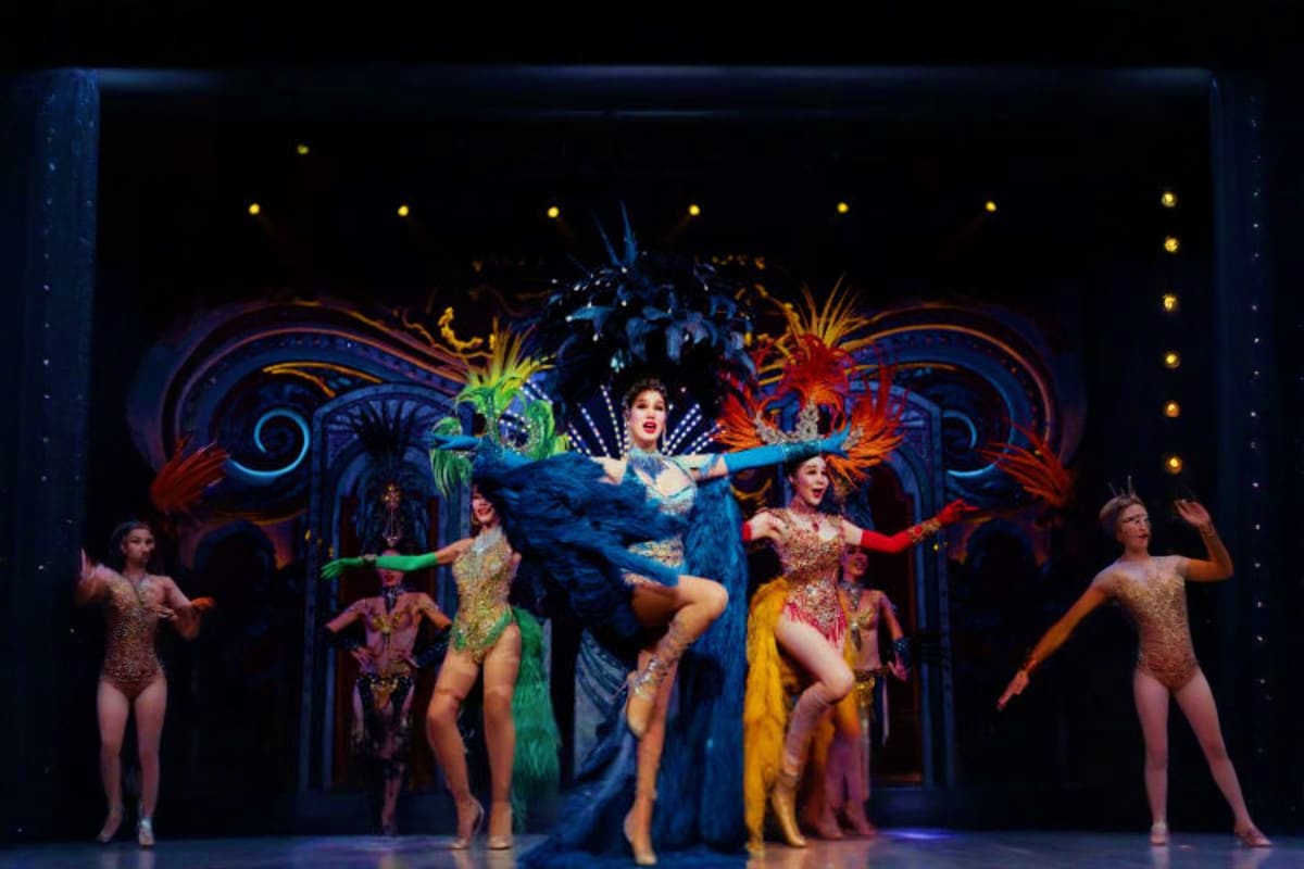 Alcazar Show performers in colorful feathered costumes dancing on stage with ornate blue backdrop and golden lights