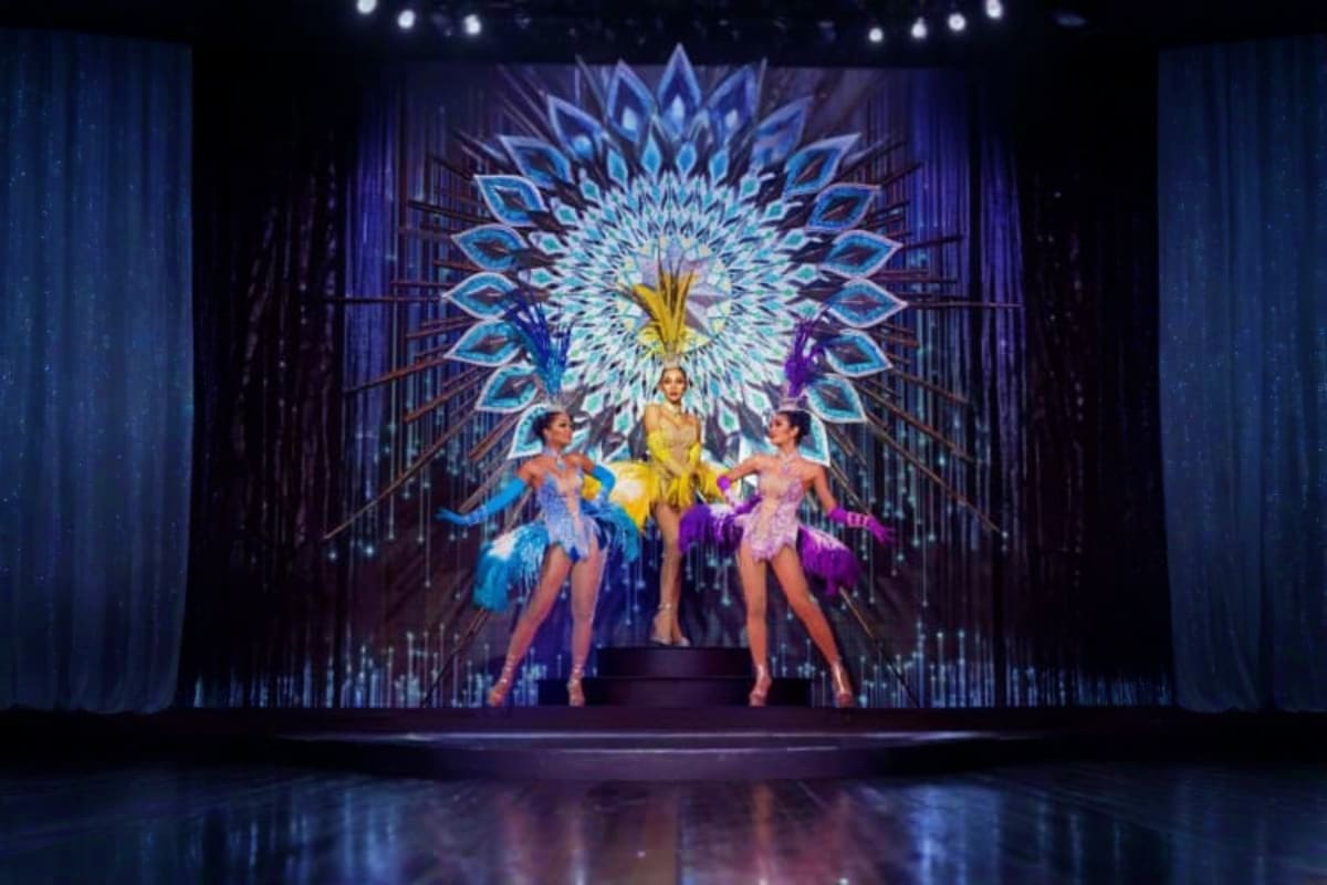 Three performers in colorful feathered costumes dancing on stage with an illuminated blue and gold peacock-inspired backdrop 