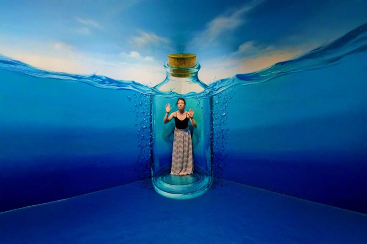 Woman standing inside a giant glass bottle submerged underwater with sky visible above the water surface