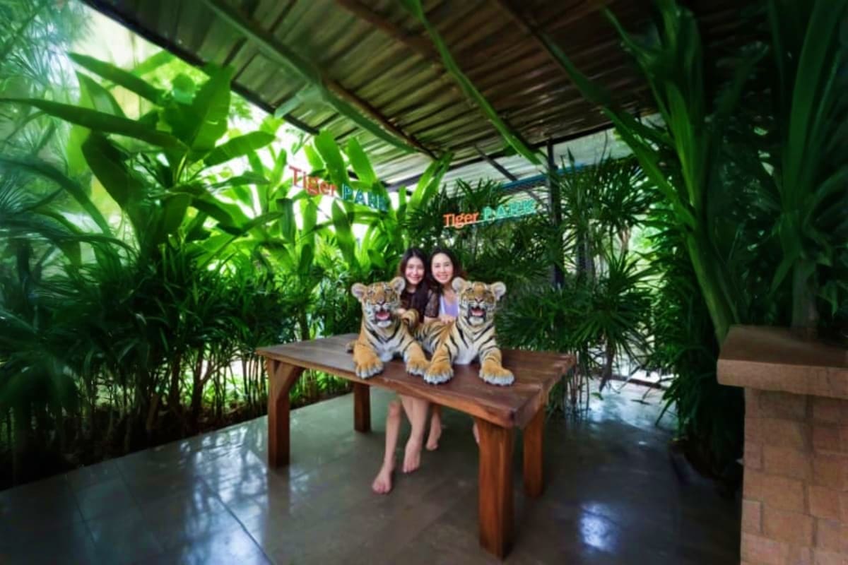 Two women posing with two tigers on a wooden table surrounded by tropical plants under a covered shelter at Tiger Park