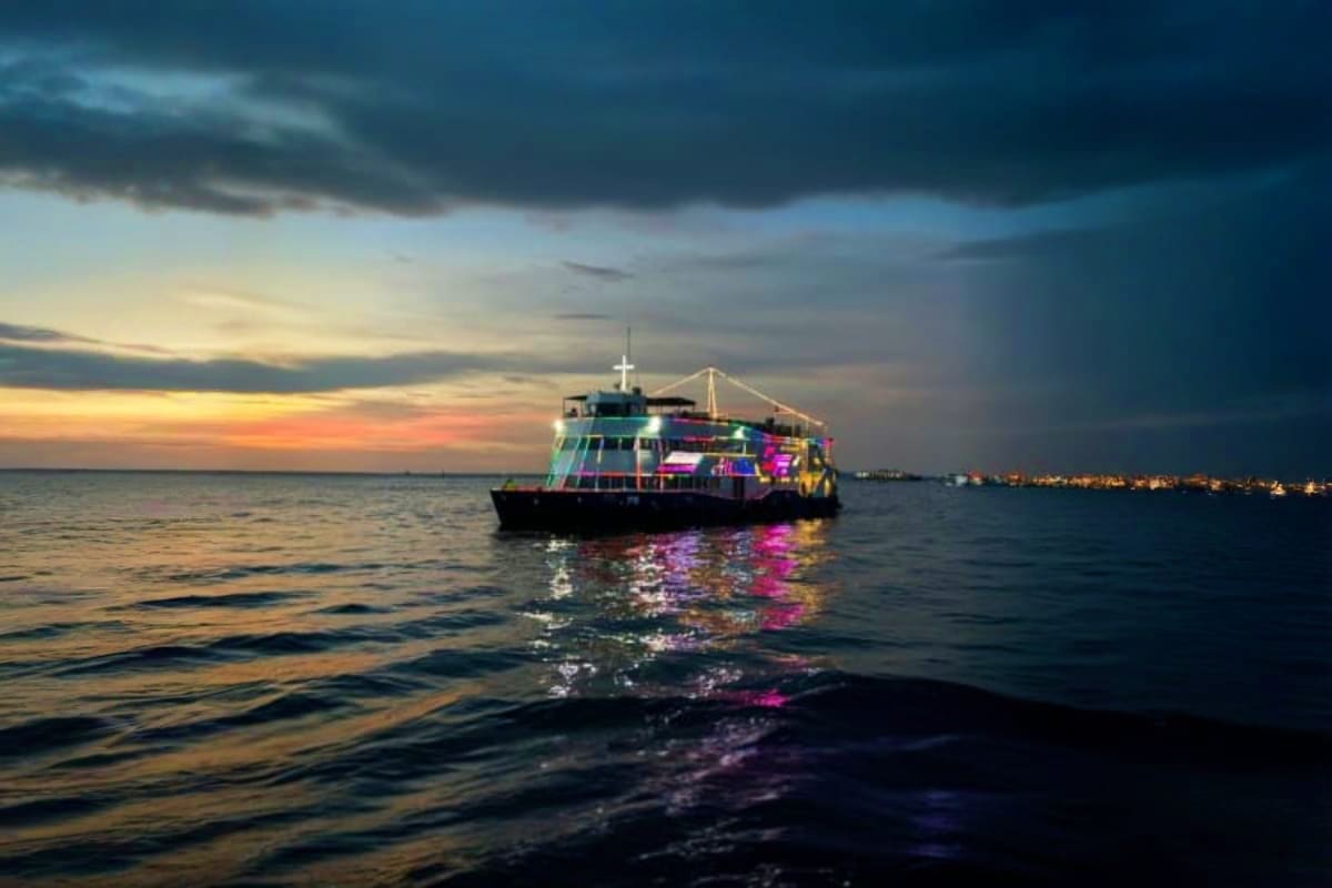 Colorful illuminated cruise ship anchored in calm ocean waters at sunset with city lights visible on the horizon