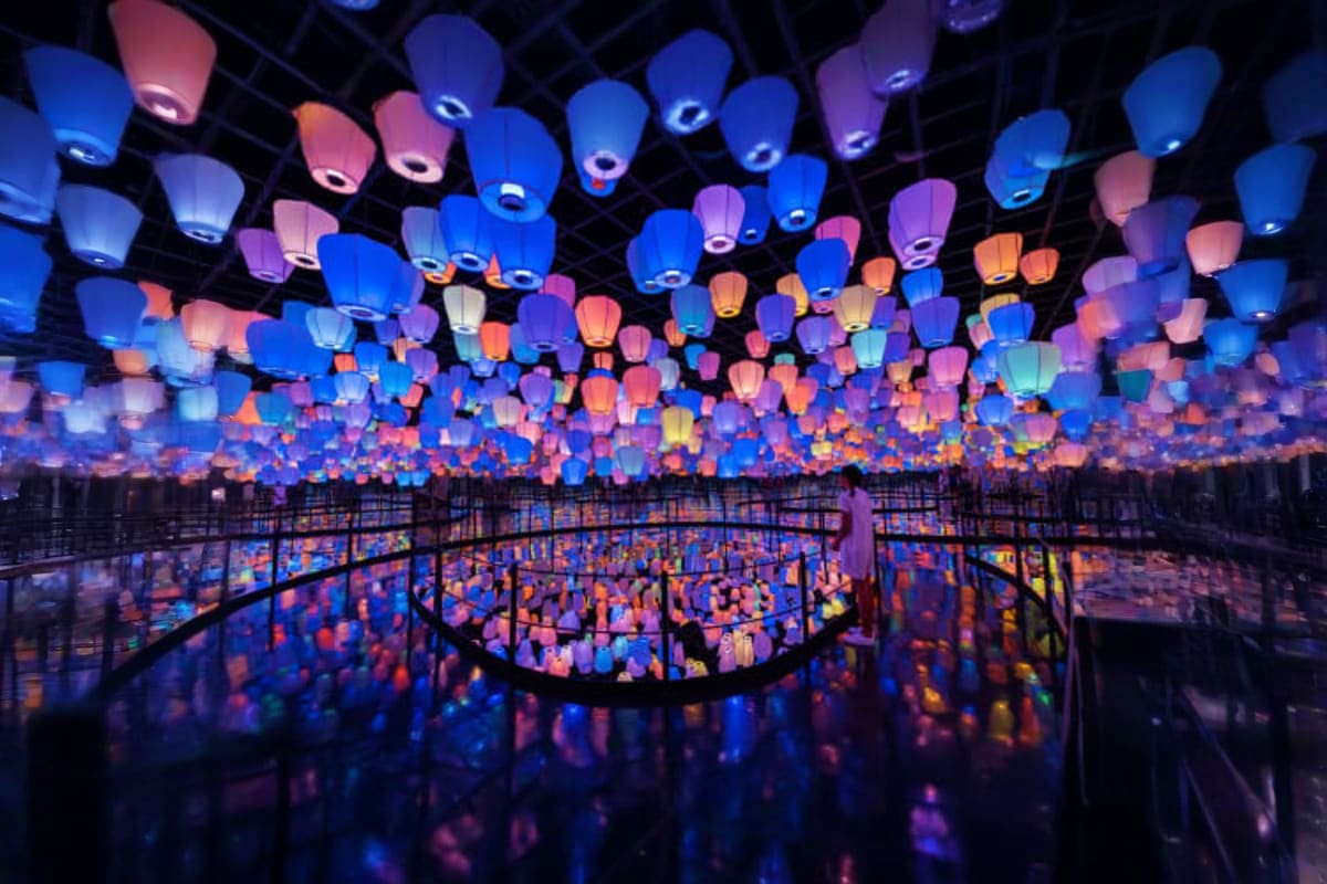 Hundreds of illuminated paper lanterns in blue, pink, and orange suspended from ceiling above reflective floor in immersive a