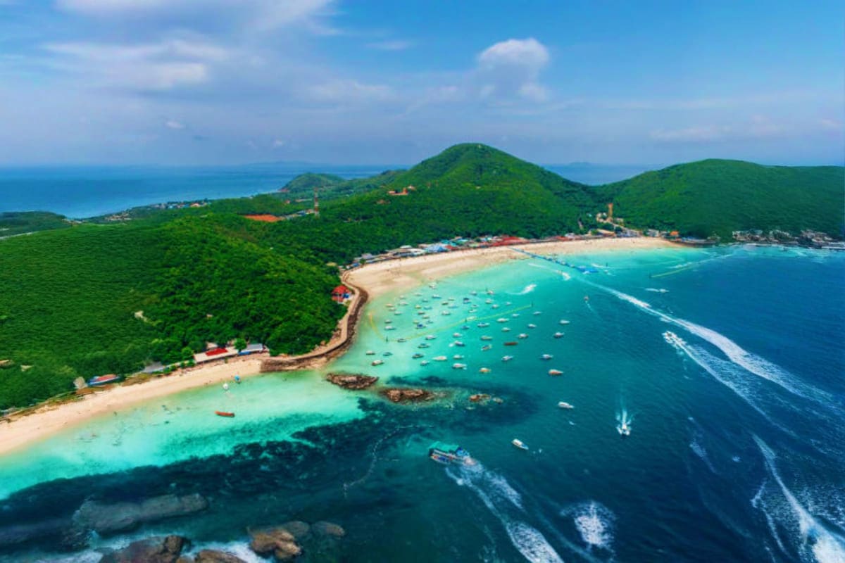 Aerial view of Coral Island beach with turquoise waters, sandy shore, forested hills, and anchored boats under blue sky