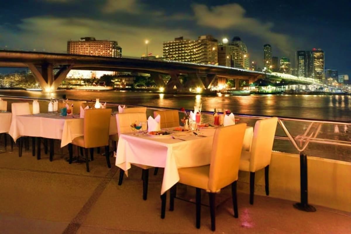 Outdoor riverfront dining tables with orange chairs and white tablecloths overlooking illuminated bridge and city skyline at 