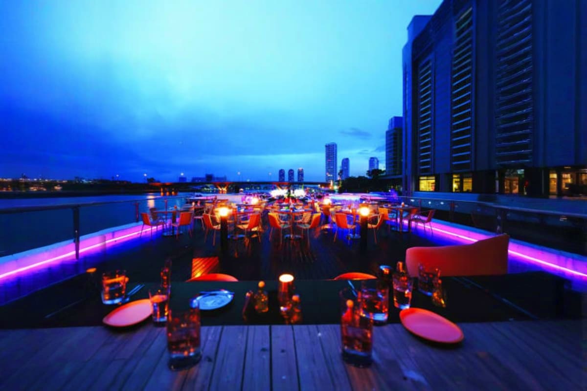 Grand Pearl Cruise Rooftop bar at twilight with neon pink and blue lighting, waterfront dining tables, city skyline, and water views