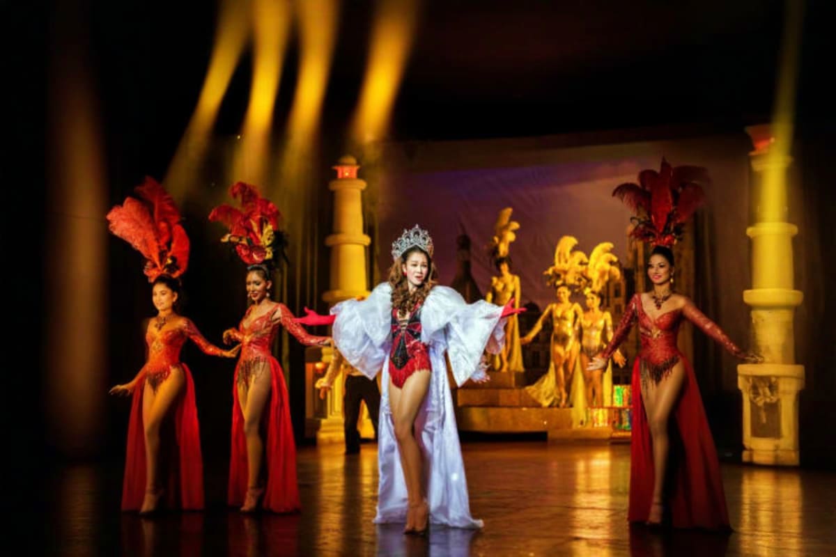 Cabaret performers on stage wearing elaborate red and white costumes with feathered headdresses, surrounded by golden archite