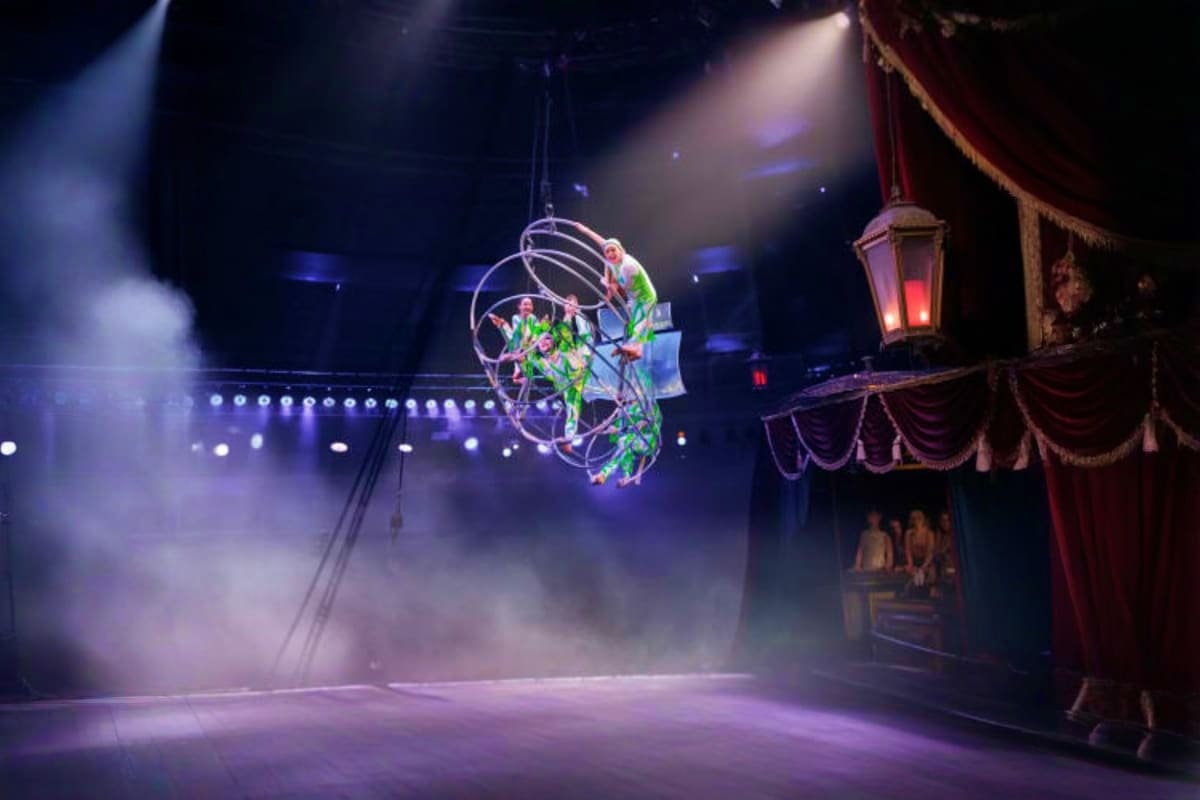 Acrobat performer suspended in aerial hoop apparatus above circus ring with dramatic lighting and red velvet stage drapes