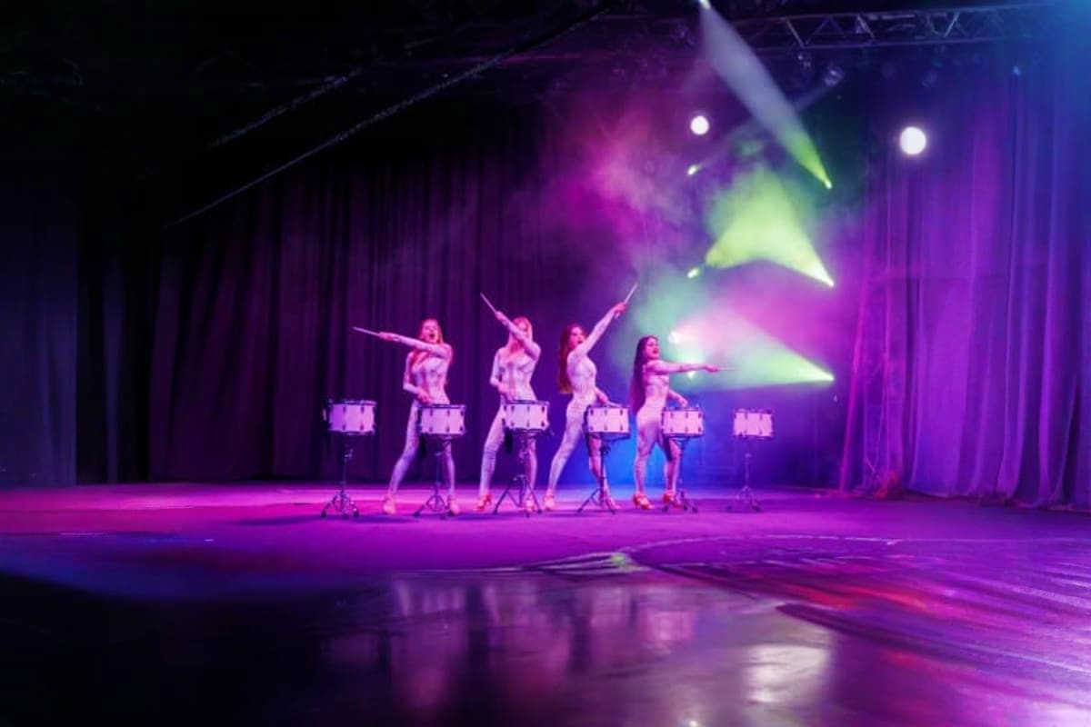 Five performers on illuminated platforms performing synchronized drum dance routine on stage with purple curtains and colorfu
