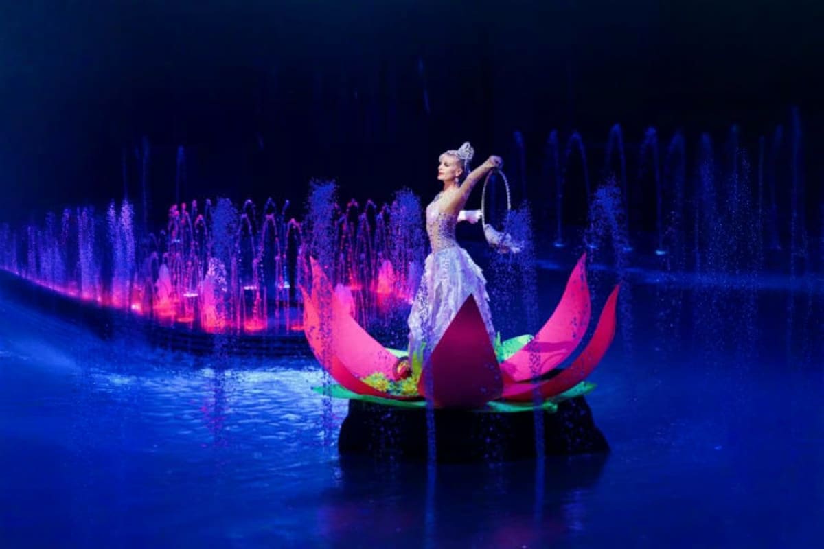Performer in white costume standing on illuminated pink boat with hula hoop during Phuket Aqua Circus water show with blue fo