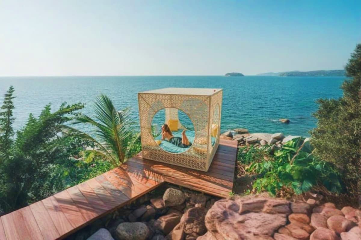Woman relaxing in woven cabana swing on wooden deck overlooking turquoise lake water and rocky shoreline with evergreen trees