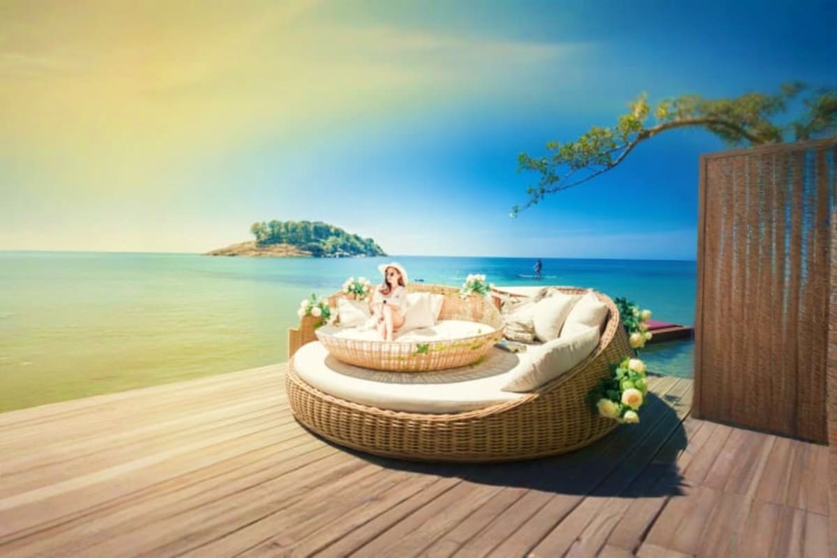 Woman relaxing in woven round daybed on wooden deck overlooking tropical beach with island and turquoise ocean