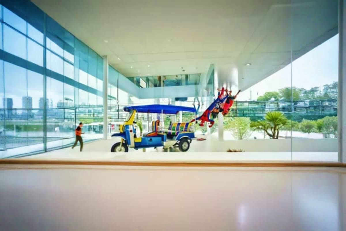 The Upside Down House Museum Modern children's play space with colorful blue and yellow playhouse structure, slide, and tuk-tuk vehicle replica in glass-w