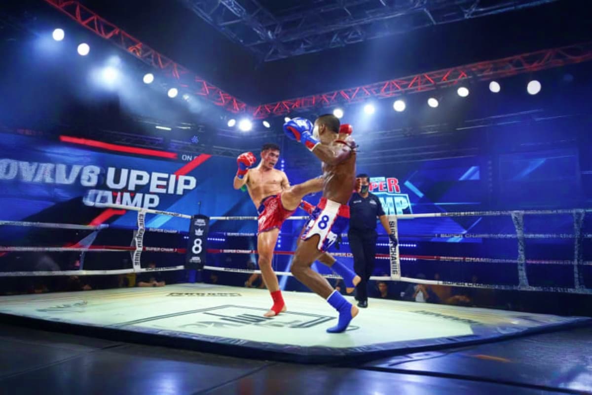 Patong Boxing Stadium Two Muay Thai fighters in red and blue shorts exchanging kicks in a brightly lit boxing ring at Patong Boxing Stadium with re
