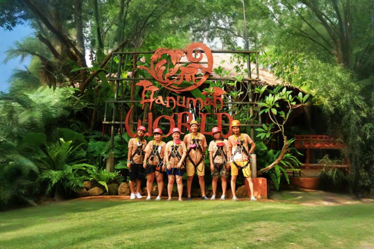 Group of six tourists in safety gear standing in front of Hanuman World zipline adventure park sign surrounded by tropical tr