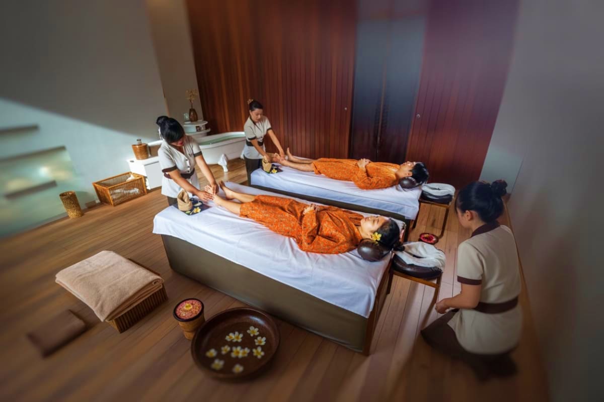 Therapists providing traditional Balinese massage treatments to two clients lying on massage beds in a spa room with wooden f